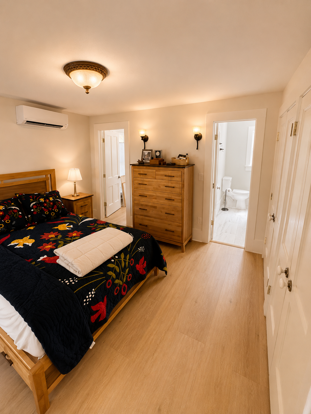 Alternate view of remodeled bedroom with light wood flooring, paneled doors, wood furniture, and adjoining bathroom.